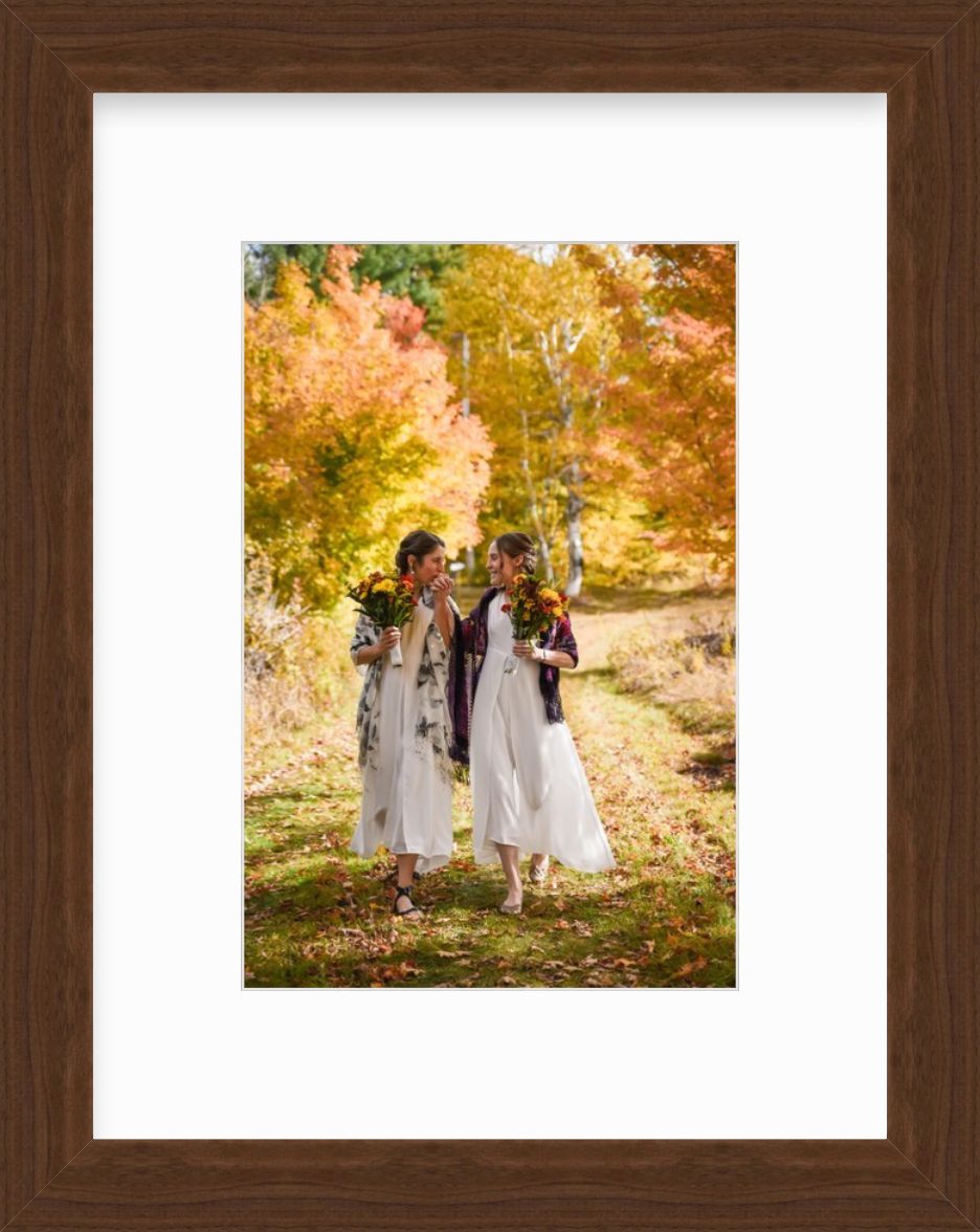Two women, just married, in white dresses and scarves hold bouquets of flowers outside in an autumnal setting.